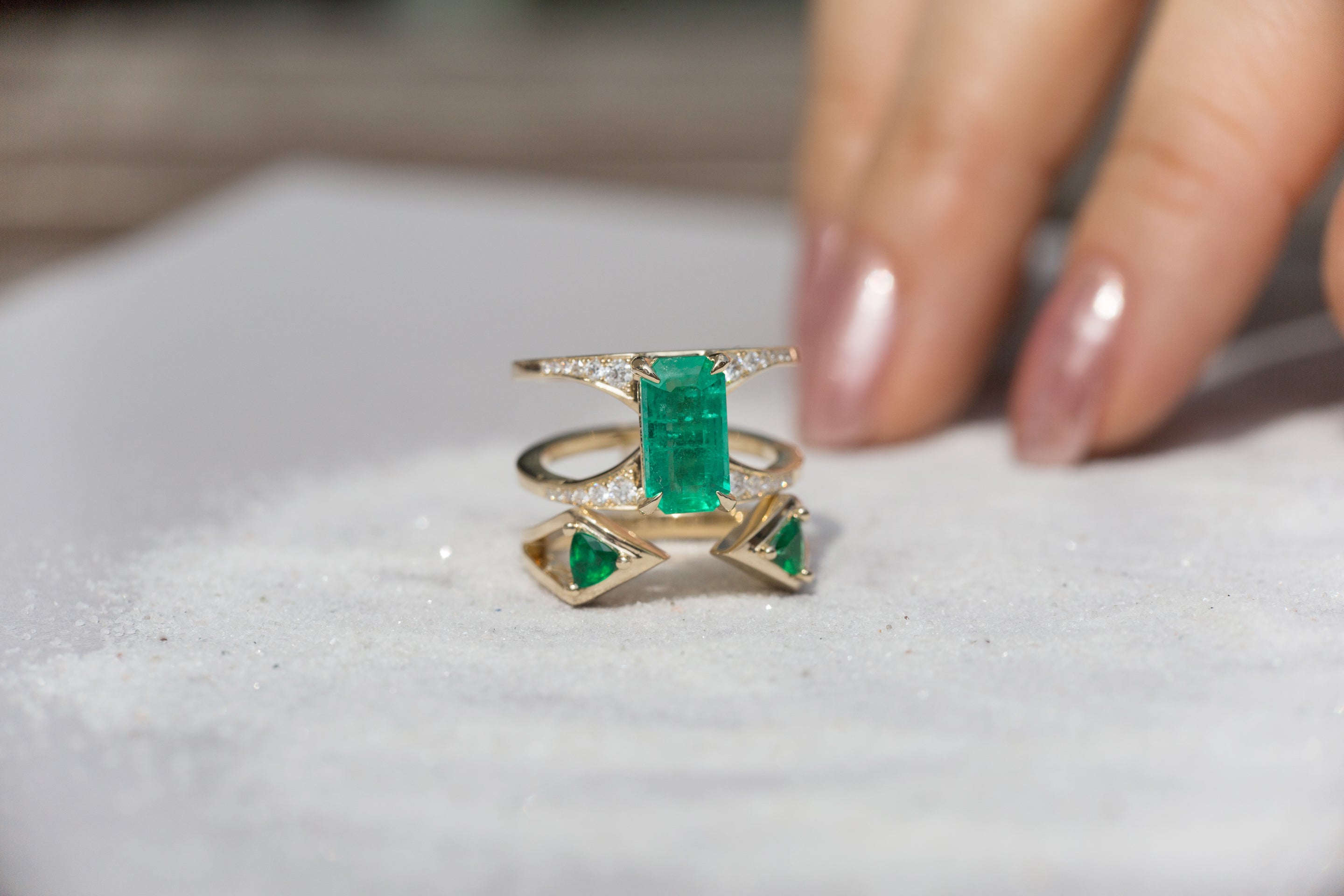 An elegant gold ring featuring vibrant emeralds, surrounded by sparkling diamonds, placed on a textured surface with a hand lightly touching it. This stunning piece showcases the beauty of emeralds.